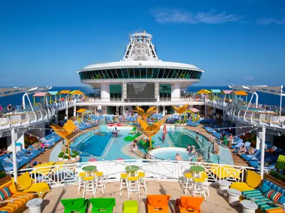 Das sonnige Pooldeck der Navigator of the Seas mit Pools, bunten Liegen und spielenden Gästen. Im Hintergrund sind das Meer und die Schiffsaufbauten zu sehen.