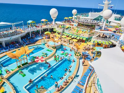 Das obere Deck der Navigator of the Seas zeigt eine weitläufige Poollandschaft mit Lazy River und vielen Gästen. Bunte Liegen und Bars säumen die Becken. Im Hintergrund das blaue Meer.