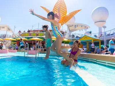 Zwei Kinder springen in den Pool auf dem Deck der Navigator of the Seas. Eine lachende Frau sitzt im Wasser. Im Hintergrund: Passagiere, Bar, Sonne.