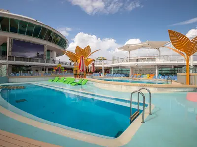 Das sonnige Pooldeck der Navigator of the Seas zeigt zwei blaue Pools, integrierte grüne Sonnenliegen, einen großen Bildschirm mit Surferbild und orangefarbene Dekorationsbäume.