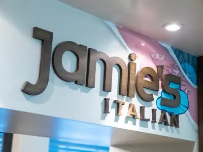 Das goldbraune 3D-Schild des Restaurants "Jamie's Italian" ist an einer Wand der Navigator of the Seas zu sehen. Die blauen Wandmalereien ergänzen das Designelement am 'S'.
