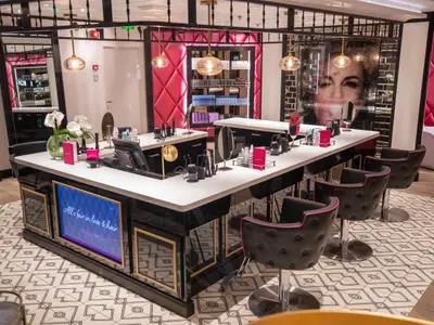 Ein stilvoller Friseursalon auf der Navigator of the Seas mit einer schwarzen U-förmigen Theke, Friseurstühlen und rosa Wänden. Schönheitsprodukte sind auf der Theke sichtbar.