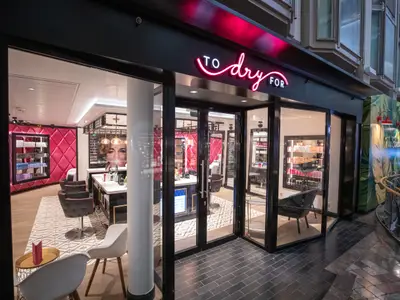 Moderner Salon 'TO dry FOR' auf der Navigator of the Seas. Durch Glasfront sichtbar: pinke Wände, Stylingstühle, Produktregale. Rechts ist der Schiffsflur erkennbar.