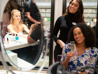 Eine lächelnde Frau mit lockigem Haar hält ein Glas Sekt. Im Hintergrund, sichtbar in einem Spiegel, erhält eine andere Frau eine Maniküre in einem hellen Salon. Zwei Crewmitglieder sind ebenfalls zu sehen.