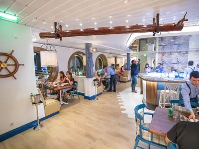 Blick in ein maritim dekoriertes Restaurant der Navigator of the Seas. Gäste speisen, Personal bedient. Im Hintergrund eine Bar mit Köchen.