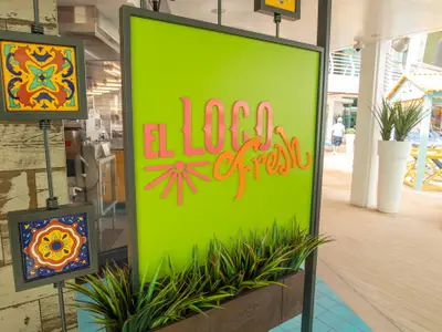 Das grüne Schild des Restaurants EL LOCO Fresh auf der Navigator of the Seas, mit pinken und orangen Lettern. Pflanzen im Vordergrund und ein Schiffsdeck im Hintergrund.