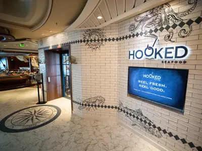 Eingangsbereich des 'Hooked Seafood' Restaurants auf der Navigator of the Seas. Weiße Kachelwände mit maritimen Illustrationen. Ein beleuchtetes Logo, ein Bildschirm und ein großes Kompassmuster auf dem Boden.