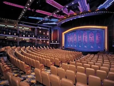 Blick in einen leeren, modernen Theatersaal an Bord der Navigator of the Seas, mit Reihen von beigen Sitzen und einer leuchtenden Bühne, die ein Kunstwerk mit Figuren zeigt. Die Decke ist mit blauer und pinker Beleuchtung versehen und es gibt mehrere Ränge mit weiteren Sitzplätzen.