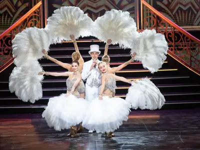 Ein Sänger in weißem Anzug und Zylinder performt mit vier Tänzerinnen in Federkostümen und Federfächern auf einer Showbühne mit Treppen im Hintergrund.