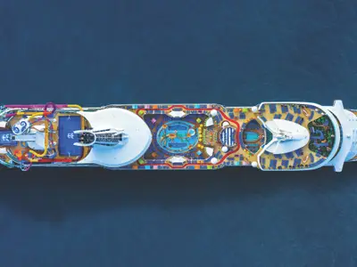 Luftaufnahme der Navigator of the Seas. Die Decks zeigen bunte Wasserrutschen, Basketballfeld, Pools und viele Liegestühle auf blauem Meer.