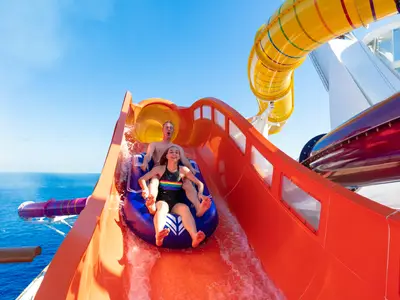 Zwei lachende Personen gleiten auf einem blauen Reifen eine orangefarbene Wasserrutsche auf der Navigator of the Seas hinunter. Im Hintergrund sind das Meer und weitere bunte Rutschen sichtbar.