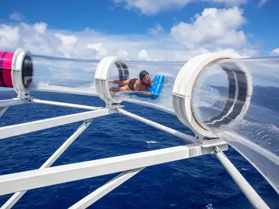 Eine junge Frau rutscht kopfüber auf einem blauen Schwimmbrett durch eine transparente Wasserrutsche. Unter ihr breitet sich das weite blaue Meer aus. Der Himmel ist blau mit weißen Wolken.
