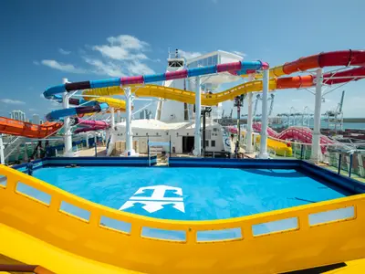 Blick auf das obere Deck der Navigator of the Seas mit vielen bunten Wasserrutschen und einem blauen Pool. Im Hintergrund sind Hafenkräne und eine Stadt zu erkennen.