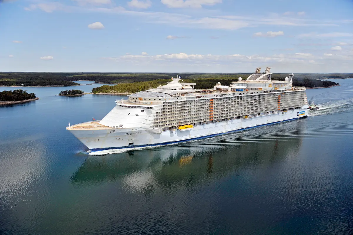 Das Megaschiff Oasis of the Seas fährt durch ein bewaldetes Schärengebiet. Ein Schlepper begleitet das Heck. Blauer Himmel mit Wolken über dem großen Schiff.