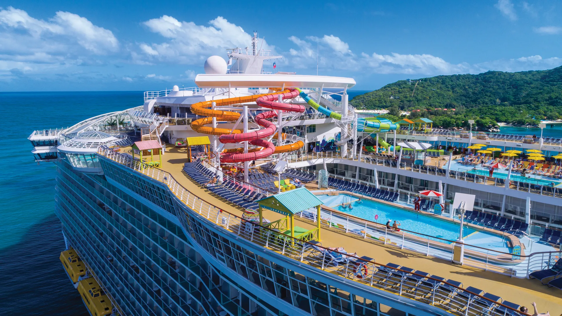 Luftaufnahme der Oasis of the Seas. Die bunten Wasserrutschen und Pools auf dem Oberdeck sind vor einer grünen Inselkulisse zu sehen.