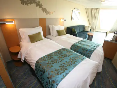 Helle Kabine auf der Oasis of the Seas mit zwei Einzelbetten, blauem Sofa, Schreibtisch und großem Fenster. Die Betten sind mit weißen Laken und dekorativen blau-goldenen Läufern versehen.