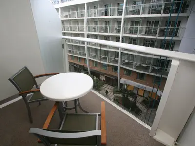 Sicht von einer Balkonkabine der Oasis of the Seas in eine mehrstöckige interne Promenade. Ein kleiner Tisch und zwei Stühle stehen auf dem Balkon. Unten sind Geschäfte und weitere Balkone zu sehen.
