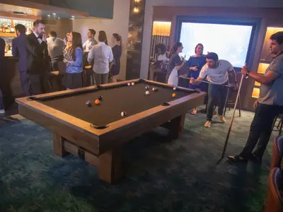 Mehrere Gäste spielen Billard in einer modernen Bar-Lounge mit dunklem Billardtisch, Bar und großer Leinwand im Hintergrund. Stimmungsvolle Beleuchtung und einige Personen unterhalten sich in kleinen Gruppen.