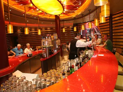 Ein Barkeeper serviert Drinks an der roten Bar der Oasis of the Seas. Mehrere Gäste sitzen an der Theke, umgeben von warmer Beleuchtung und stilvoller Einrichtung.