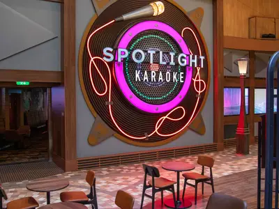 Karaoke-Lounge 'Spotlight Karaoke' auf der Oasis of the Seas. Großes Neon-Schild mit Mikrofon und Schriftzug. Sitzgelegenheiten im Vordergrund.