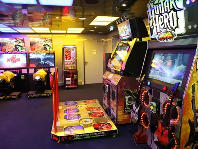 Eine moderne Spielhalle auf der Oasis of the Seas mit Racing-Automaten, einem Tanzspiel und Guitar Hero. Bunte Beleuchtung erhellt den Raum an Bord.