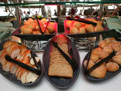 Brotbuffet an Bord der Oasis of the Seas: Eine Auswahl an frischen Brötchen, geschnittenem Baguette und Vollkornbrot in Servierschalen mit Zangen. Gäste und weitere Buffetbereiche im Hintergrund.