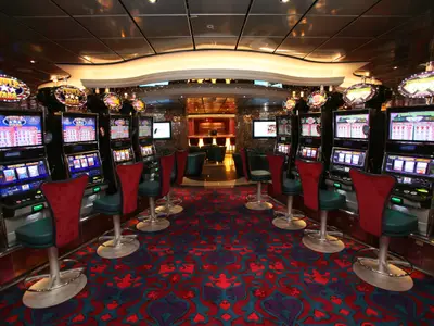 Das Casino der Oasis of the Seas zeigt Spielautomaten mit roten und türkisen Barhockern. Der Boden ist mit gemustertem Teppich bedeckt, im Hintergrund ist eine Bar zu sehen.