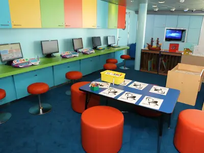 Der helle Kinderbereich an Bord der Oasis of the Seas zeigt eine Computerstation mit farbigen Tastaturen, einen Maltisch mit orangen Sitzgelegenheiten und Regale mit Büchern und Spielzeug.