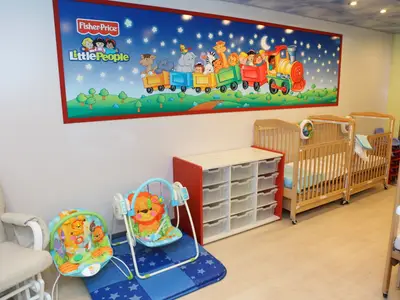 Ein heller und freundlicher Baby- und Kleinkindbereich an Bord, ausgestattet mit Holzkrippen, einem Wickeltisch, Babyschaukeln und einer Spielmatte. Eine große Wanddekoration mit einem Cartoon-Zug und Tieren ziert den Raum.