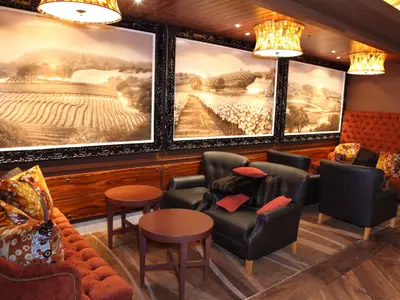 Innenansicht einer Lounge auf der Oasis of the Seas. Weinbergsbilder, Ledersessel, Sofas und Holztische sind zu sehen. Besondere Deckenleuchten erhellen den Raum.