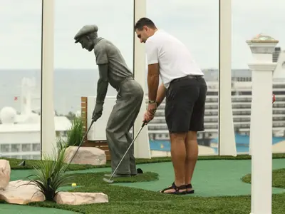 Ein Mann und eine Statue spielen Minigolf auf dem Oberdeck der Oasis of the Seas. Im Hintergrund sind Meer und Schiffsaufbauten durch große Fenster sichtbar.