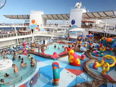 Das Pooldeck der Oasis of the Seas. Badegäste in Pools und auf einem farbenfrohen Wasserspielplatz mit bunten Elementen. Sonniger Tag.