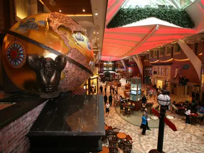 Panorama Royal Promenade der Oasis of the Seas. Goldene Weltkugel mit Atlas-Statue prominent. Dahinter belebte Geschäfte, Cafés und Passagiere unter rotem Dach.