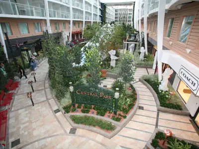 Der Central Park an Bord der Oasis of the Seas. Üppige Grünflächen, Spazierwege, Geschäfte wie COACH und Skulpturen sind sichtbar. Der Park wird von Balkonkabinen eingerahmt.