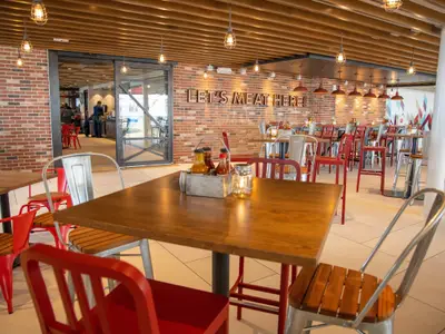 Innenansicht eines modernen Restaurants mit Holztischen, roten und silbernen Stühlen. Eine Backsteinwand trägt den leuchtenden Schriftzug 'LET'S MEAT HERE!'. Im Vordergrund stehen Gewürzflaschen und ein Kerzenglas auf einem Holztisch. Im Hintergrund sind weitere Tische und eine Tür zu sehen, dahinter Mitarbeiter.