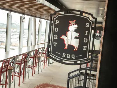Die 'Port Side BBQ' Bar auf der Oasis of the Seas. Eine lange Theke mit Barhockern, große Fenster mit Hafenblick. Markantes Logo mit gestapelten Tiermotiven.