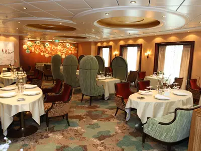 Innenansicht eines eleganten Restaurants auf der Oasis of the Seas mit gedeckten Tischen, grünen Hochlehnern und roten Sesseln, umgeben von geschmackvoller Wandkunst und großen Fenstern.