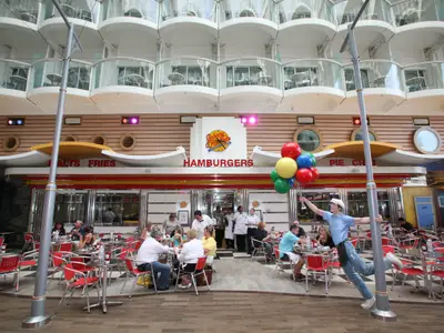 Das Johnny Rockets Diner auf der Oasis of the Seas mit sitzenden Gästen und Personal. Ein Entertainer hält bunte Ballons. Im Hintergrund sind die Schiffsbalkone sichtbar.