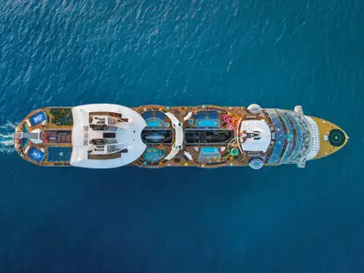 Luftaufnahme der Oasis of the Seas auf blauem Meer. Bug, Heck mit Heckwelle, Pools, Rutschen, Sportplätze und ein Helipad am Bug sind sichtbar.