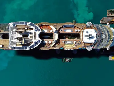 Luftbild der Oasis of the Seas im Hafen. Man sieht die Oberdecks mit Pools, Gärten und Freizeitanlagen. Kleine Boote neben der Oasis of the Seas im türkisblauen Wasser.