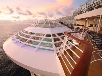 Blick auf das Oberdeck der Oasis of the Seas im Abendlicht. Ein Glasdom über einem Pool ist zentral, umgeben von Reling und Sonnenliegen, mit dem Meer im Hintergrund.