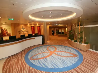 Der Empfang des Vitality at Sea Spa & Fitness Centers auf der Oasis of the Seas. Mit Theke, Holzvertäfelung, rundem Teppich und Pflanzen. Gang führt zum Spa.