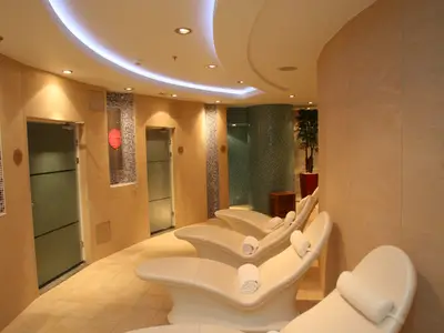 Ein heller und moderner Spa-Bereich mit mehreren cremefarbenen Entspannungsliegen, warmem Licht und hellen Fliesenwänden. Links sind Türen zu sehen, mittig eine Wand mit Mosaikfliesen und einem runden roten Dekorationselement. Rechts hinten ist eine Grünpflanze in einem roten Topf.