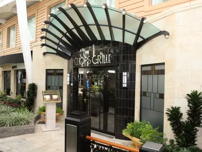 Detailansicht des Eingangs zum Restaurant Chops Grille mit glänzend schwarzen Fliesen und einem geschwungenen Glasvordach. Links und rechts der Tür befinden sich schlanke Pflanzenbeete und ein beleuchtetes Menüpult. Die Fenster links zeigen weitere Räumlichkeiten. Der obere Gebäudeteil ist hell sandfarben.