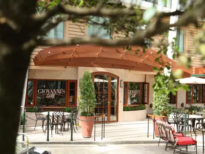 Die Fassade des Restaurants 'Giovanni's Table' mit gewölbtem Holzdach, großen Fenstern und Außenbereich. Zahlreiche Tische und Stühle, umgeben von Pflanzen und Zäunen.