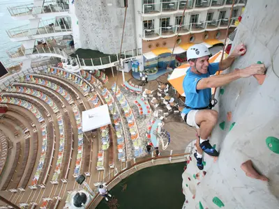 Ein lächelnder Mann klettert an der Wand auf der Oasis of the Seas. Im Hintergrund: leere Sitzreihen, Kabinen, Meer.