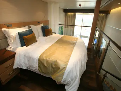 Blick auf den Schlafbereich einer Loft-Suite der Oasis of the Seas. Großes Bett, dahinter Glasgeländer zur unteren Ebene mit Fensterfront.