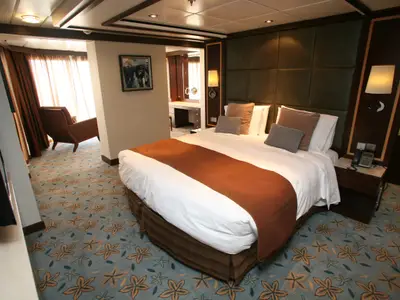 Komfortable Kabine auf der Oasis of the Seas. Großes Bett, Sessel am Fenster, Schminktisch. Blauer Teppich mit maritimen Mustern.
