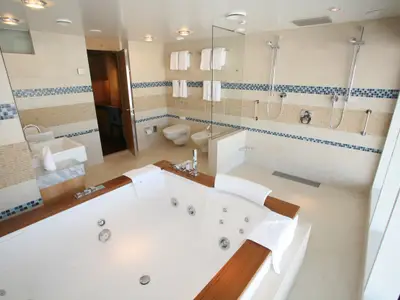 Ein geräumiges Badezimmer auf der Oasis of the Seas. Es zeigt einen großen Whirlpool, eine Doppel-Dusche, ein WC, ein Bidet und ein Waschbecken. Die Wände sind hell gefliest mit blauen und beigen Mosaikbändern.