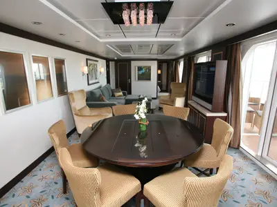 Innenansicht einer luxuriösen Suite der Oasis of the Seas. Das Bild zeigt einen Wohnbereich mit Esstisch, bequemen Möbeln, einem großen TV und Zugang zu einem Balkon mit Sitzgelegenheiten.